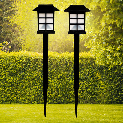 Two black solar garden lights with white paneled tops stand in a vibrant green lawn with hedges and trees.